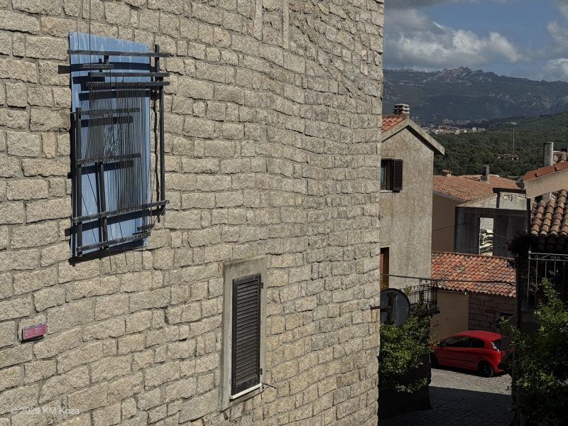 Photo of Aggius buildings with artwork loom on building and in distance, mountains.